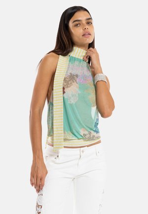 Vrouw draagt een mouwloze bloemen top met geel gestreepte sjaaldetail, witte broek met kanten uitsnijdingen en een witte kralen armband, poseert tegen een witte achtergrond.