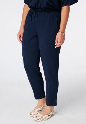 Vrouw draagt marineblauwe taps toelopende broek met trekkoord in de taille, gecombineerd met beige flats met rasterpatroon en gouden armbanden aan haar rechterpols.