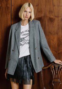Donna bionda che indossa un blazer grigio sopra una maglietta bianca con grafica e una gonna in pelle nera con frange, appoggiata su una sedia in legno intagliata contro una parete di pannelli di legno.