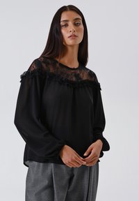 Giovane donna con lunghi capelli scuri che indossa una blusa nera trasparente con dettagli in pizzo e pantaloni grigi su misura, contro uno sfondo semplice.
