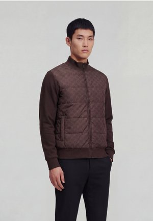 Ung mand iført en brun quiltet jakke med lynlås og sorte bukser, stående med den ene hånd i lommen mod en ensfarvet baggrund.