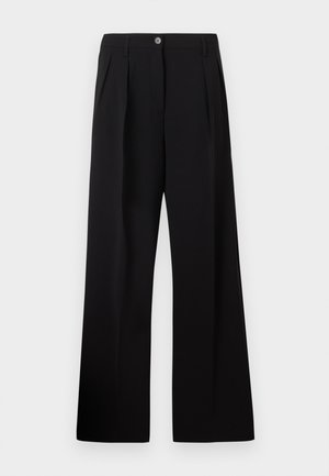 Γυναίκα στέκεται φορώντας λευκό πουκάμισο με κουμπιά, μαύρο φαρδύ παντελόνι με ψηλή μέση και μαύρα μυτερά φλατ παπούτσια με διακοσμητικά αγκράφες.