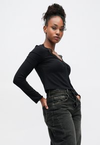 Černý top s dlouhými rukávy a krajkovým lemováním kolem výstřihu a manžet, doplněný vysokým pasem černých džínsů s viditelným prošíváním.