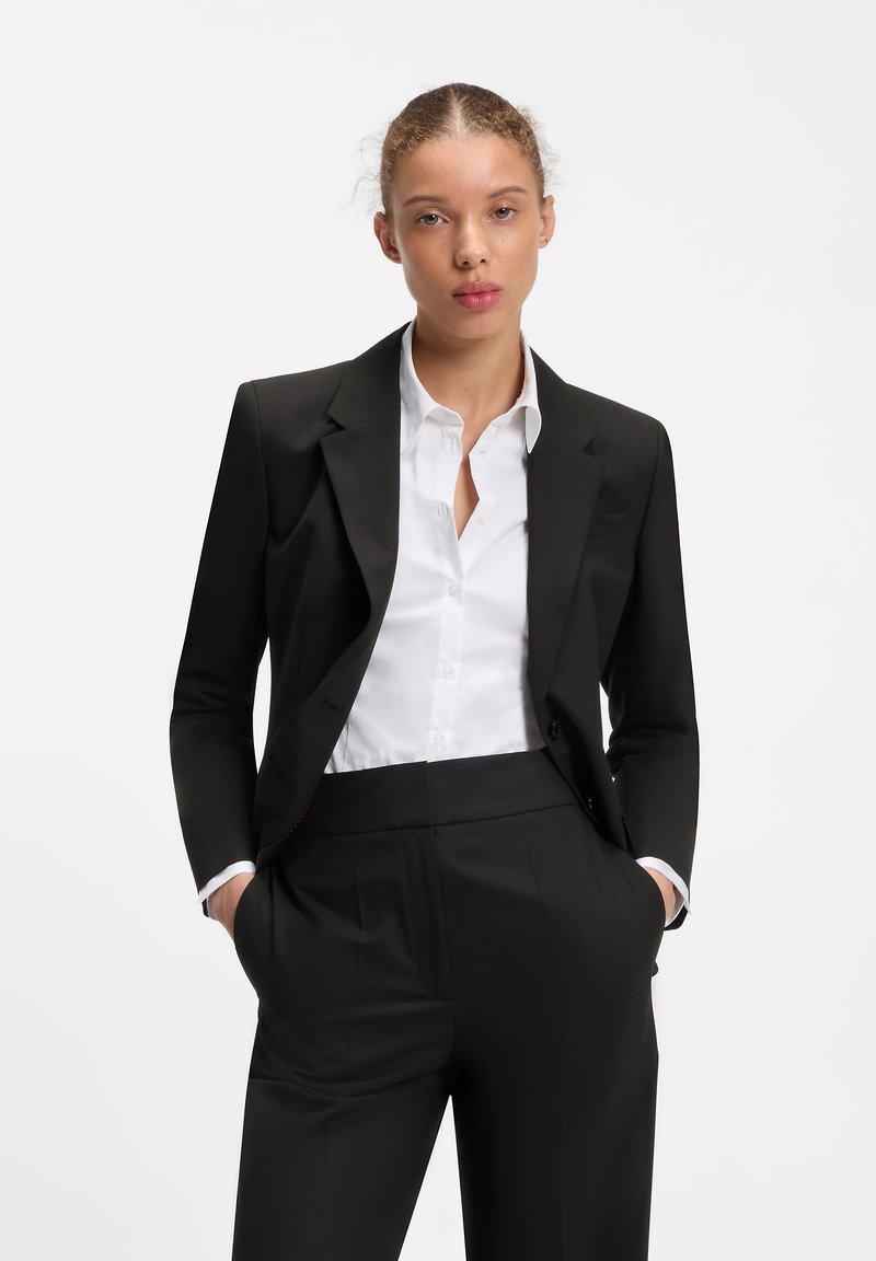 Jeune femme aux cheveux attachés, portant une veste et un pantalon noirs avec une chemise blanche boutonnée, les mains dans les poches, debout devant un fond blanc.