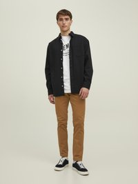 Czarna koszula z guzikami założona na białą koszulkę z grafiką, w połączeniu z brązowymi spodniami slim fit i granatowymi sneakersami. Prosty design i neutralne kolory.