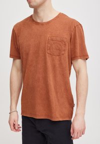 Camiseta de algodón de color óxido con mangas cortas, cuello redondo y un bolsillo en el pecho. Textura suave con una apariencia sutilmente desvanecida y dobladillo cosido.