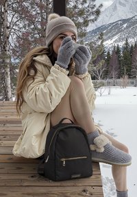 Zaino nero con zip dorata, pantofole beige in peluche, guanti grigi a costine e una giacca a vento crema, posizionati davanti a alberi e montagne innevati.