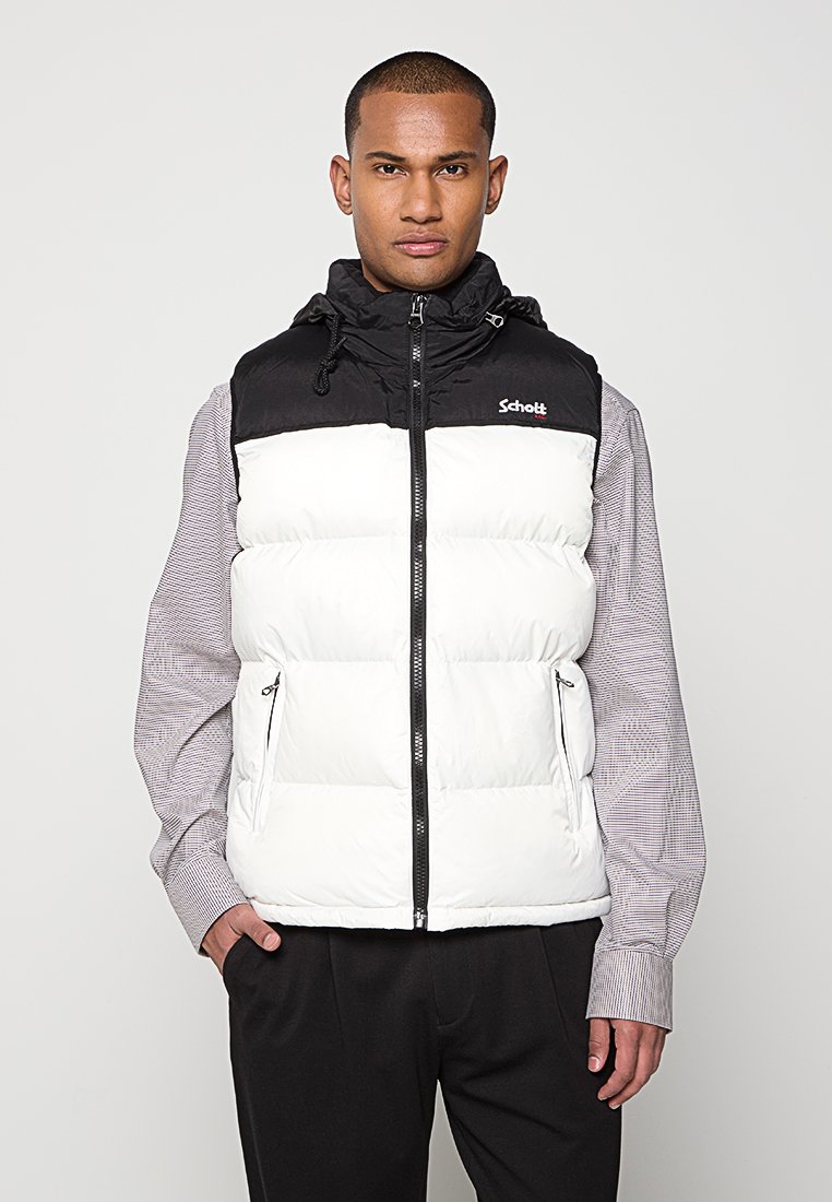 Homme portant un gilet rembourré sans manches noir et blanc avec capuche sur une chemise à manches longues à carreaux et un pantalon noir, se tenant devant un fond uni.
