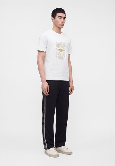Νέος άνδρας που φοράει λευκό t-shirt Emporio Armani, μαύρο παντελόνι με λευκές πλαϊνές ρίγες και μπεζ αθλητικά παπούτσια, στέκεται μπροστά σε απλό φόντο.