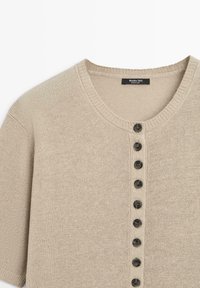Beige gebreide trui met korte mouwen, een ronde hals en een verticale rij van acht donkere knopen in het midden vooraan.