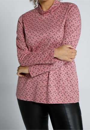 Femme croisant les bras, portant un haut rose à manches longues avec de petits motifs floraux noirs et un pantalon en cuir noir, sur un fond uni.