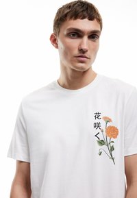 Camiseta de algodón blanca con gráfico floral en naranja y verde, con texto negro en el lado izquierdo. Manga corta y cuello redondo.