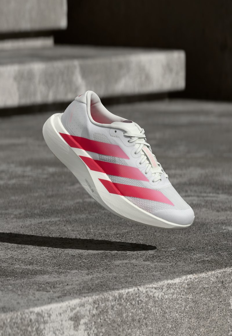 Scarpa da corsa bianca con tre grintose strisce rosse, sospesa in aria sopra gradini di cemento, che mostra un design leggero e suola testurizzata.