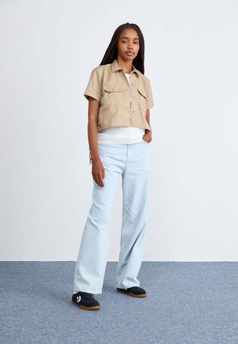 Camisa beige recortada de manga corta con dos bolsillos en el pecho, combinada con jeans de pierna ancha azul claro y zapatillas negras. Fondo liso.