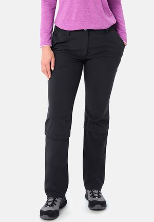 Vrouw draagt zwarte ombouwbare wandelbroek, paarse longsleeve en grijze wandelschoenen, staat met handen in de zakken.