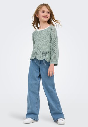 Young girl smiling, wearing a mint green knit sweater, wide-leg blue jeans, and white sneakers, standing against a plain background.