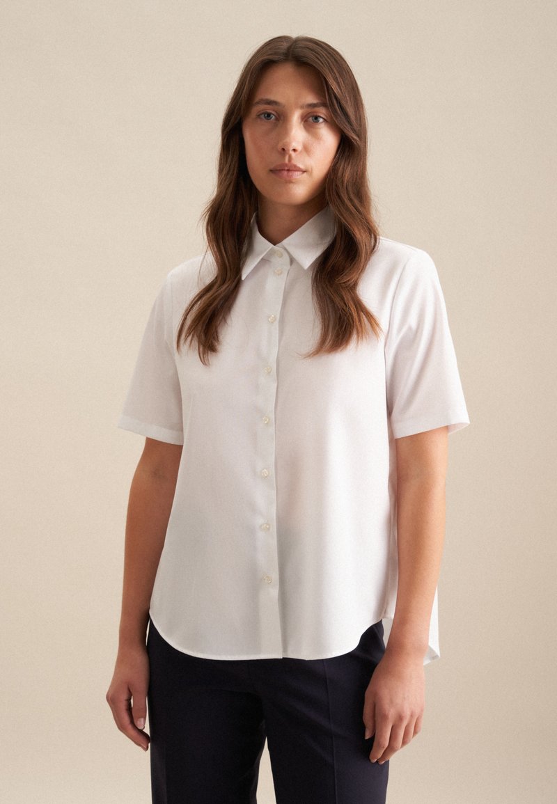 Camicia bianca a maniche corte, con texture liscia, colletto classico e orlo curvo, abbinata a pantaloni scuri su uno sfondo neutro.