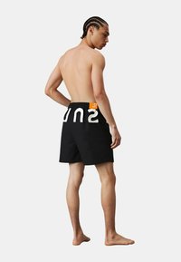 Pantaloni da bagno neri con lettering bianco "SUN", un'etichetta arancione sulla vita e un materiale leggero, mostrati dal retro.