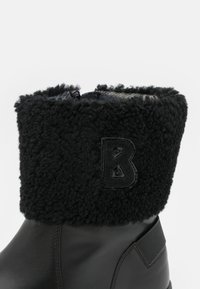 Czarne skórzane buty z teksturowanym czarnym kołnierzem z futra, wytłoczonym logo "B" i gładkim wykończeniem. Akcentowane kontrastującą wkładką.