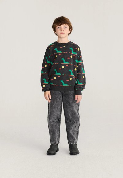 Garçon portant un pull sombre avec des motifs de dinosaures verts, un jean gris ample et des bottes noires, debout devant un fond clair uni.