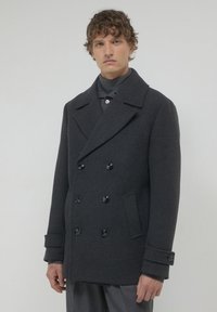 Young man with curly hair wearing a dark gray double-breasted pea coat and gray trousers against a plain background.