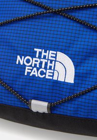 Modra teksturirana tkanina z izstopajočim belim vezenim logotipom The North Face, z črnim elastičnim kablom preko površine.