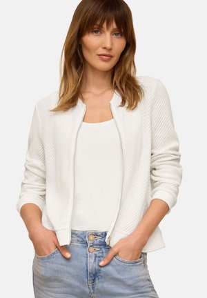 Donna con capelli castano chiaro indossa una giacca bianca traforata sopra un top bianco e jeans azzurri, mani nelle tasche, sfondo neutro.