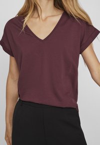 Burgunderfarbendes V-Ausschnitt-T-Shirt mit kurzen, umgeschlagenen Ärmeln und lockerem Schnitt, aus weichem Stoff gefertigt. Kombiniert mit schwarzen Hosen.