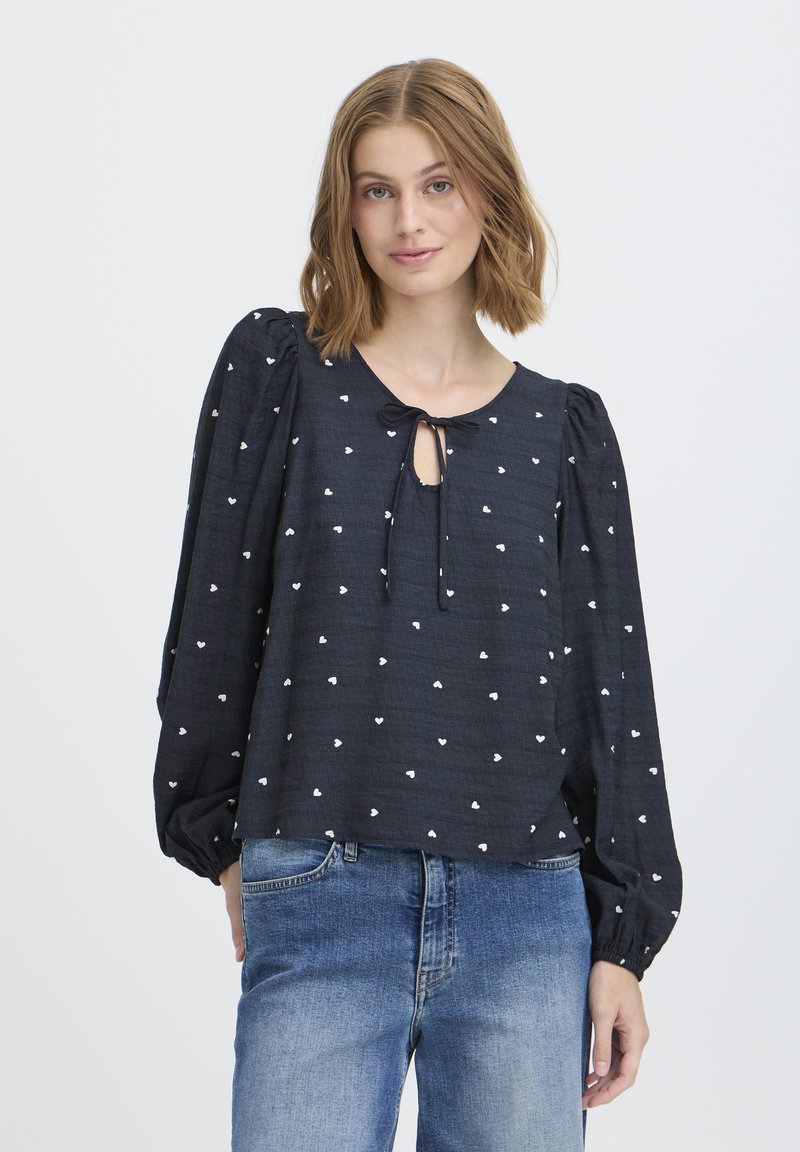  blusa de color navy con patrones de corazones blancos, escote redondo y detalle de lazo. Cuenta con mangas abullonadas y un corte holgado, fabricada con tela ligera.