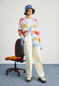 Cardigan à carreaux coloré dans des teintes pastel avec des finitions bleues, pantalons larges crème, et chaussures à plateforme beiges. Chapeau seau en denim bleu.
