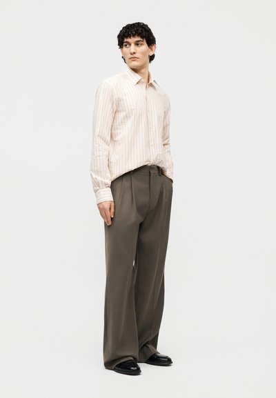 Άνδρας που φοράει ανοιχτό μπεζ πουκάμισο με λεπτές κόκκινες ρίγες, φαρδιά καφέ παντελόνια και μαύρα παπούτσια, στέκεται με τα χέρια στις τσέπες μπροστά σε λευκό φόντο.