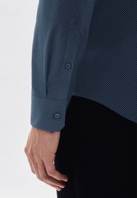 Granatowa koszula w białe kropki, z guzikami przy mankietach i gładką fakturą. Zbliżenie na rękaw i dłoń.