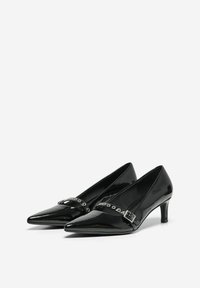Talons aiguilles en cuir verni noir avec une sangle à la cheville, présentant une boucle en argent et des détails en œillet, sur fond blanc.