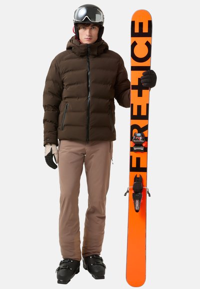 Brown puffer jacket, tan ski pants, black gloves, and ski boots. Holding an orange ski with black text. Accessories include a helmet and goggles.