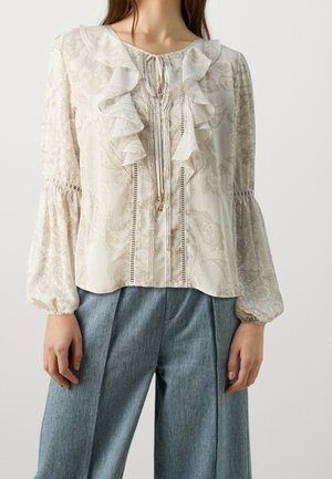 Femme portant un chemisier crème à motifs floraux avec un devant à volants et des manches ballon, associé à un pantalon en jean bleu à jambes larges.