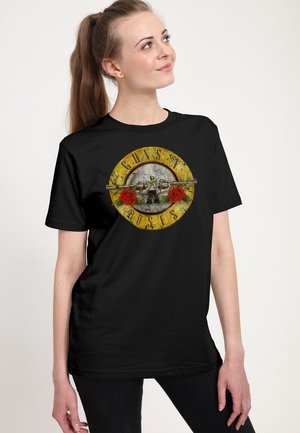 Femme portant un t-shirt noir Guns N' Roses avec le logo du groupe comportant deux roses rouges et des pistolets croisés sur un fond circulaire jaune.