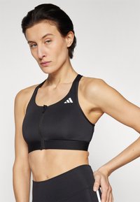 Soutien-gorge de sport noir avec une fermeture éclair à l'avant, des bretelles larges et des accents de logo blanc. Matière lisse, design ajusté, adapté aux activités sportives.