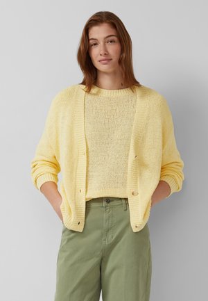 Junge Frau mit schulterlangem roten Haar, trägt einen hellgelben Strickpullover und eine passende Strickjacke sowie olivgrüne Hose, die Hände in den Taschen.