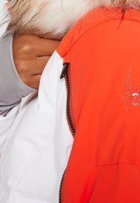 Personne portant une veste blanche et orange avec une capuche doublée de fourrure, une fermeture éclair visible et un petit logo d'araignée sur le tissu orange.