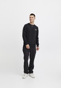 Svart sweatshirt med ribbade muddar och nederkant, utrustad med en liten logopatch på bröstet; kombinerad med lösa svarta byxor och vita sneakers.