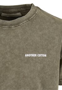 Olivově zelené bavlněné tričko s kulatým výstřihem. Na přední straně má bílý potisk s nápisem „ANOTHER COTTON“. Měkká, texturovaná látka.