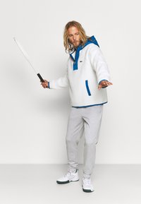 Fehér bolyhos pulóver kék kapucnival és díszítésekkel, szürke melegítőnadrággal és fehér tornacipővel párosítva. A modell egy ütővel pózol.