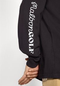 Black long-sleeve shirt featuring large white "salobongOLF" text along the arm and a small logo tag at the hem. Soft, cotton material.