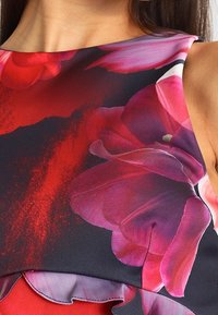 Primo piano di un abito senza maniche con stampa floreale rossa e rosa su sfondo scuro, indossato da una persona con lunghi capelli scuri.