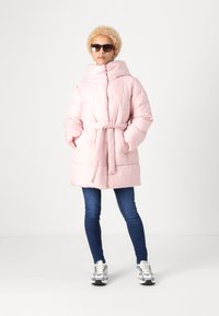 Giacca puffa rosa con ampio colletto, vita cintrata e motivo trapuntato. Indossata sopra jeans scuri e abbinata a sneakers bianche e occhiali da sole.