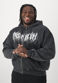 Juodas užtrauktukas džemperis su minkšta tekstūra, pasižymintis balta ugnies pavidalo grafika ant krūtinės ir kengūros kišenėmis priekyje.