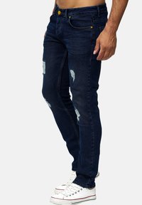 Mørkeblå skinny jeans med slidte områder foran, med gul knap og lommer. Sammen med hvide sneakers.
