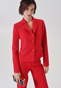 Blazer rosso su misura con due bottoni e rever a incavo, abbinato a pantaloni coordinati. Il modello tiene in mano una clutch nera con cornice dorata.