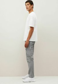 Muž v bílém oversized tričku, šedých cargo kalhotách s kapsami na klopě a bílých teniskách stojící před jednobarevným béžovým pozadím.