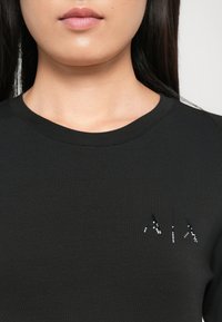 Schwarzes Baumwoll-T-Shirt mit Rundhalsausschnitt, verziert mit Strassbuchstaben "A|A|X" auf der linken Seite der Brust. Glatte Textur.